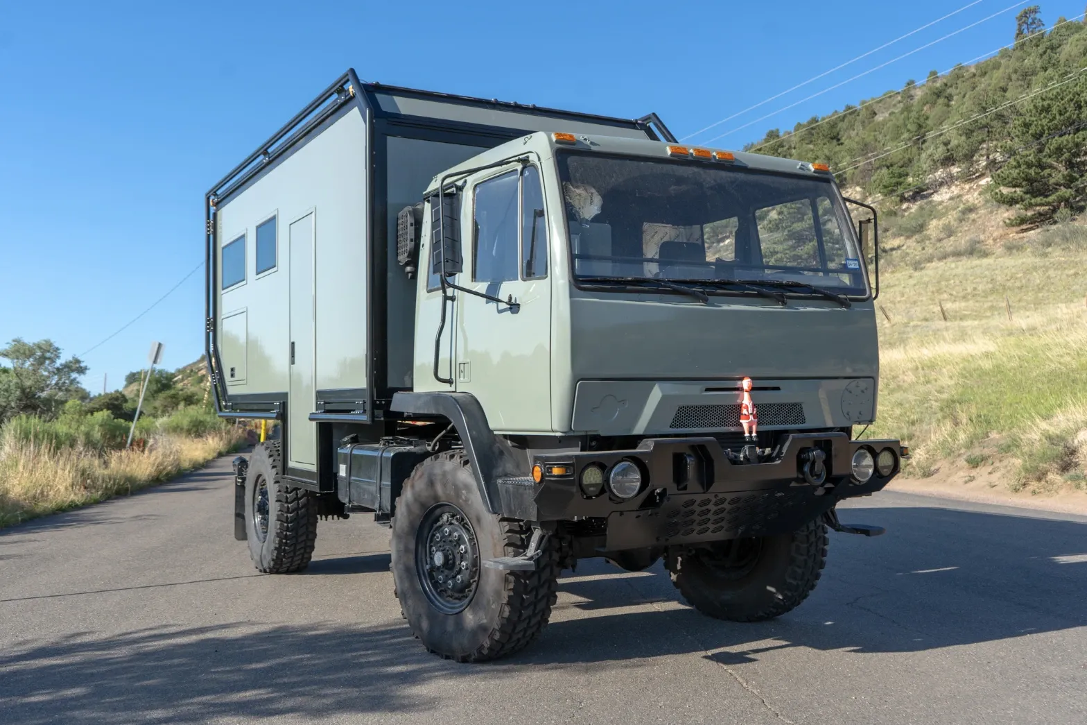 Green LMTV expedition truck with a composite camper body on a mountain road