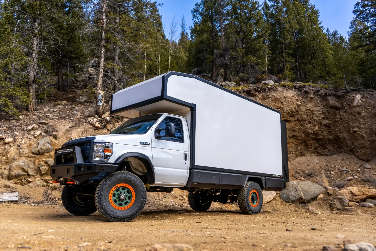 White Ford E-450 4x4 E-Series composite box camper parked on a forest dirt road, front three-quarter view showing the cab, off-road bumper and beadlock wheels