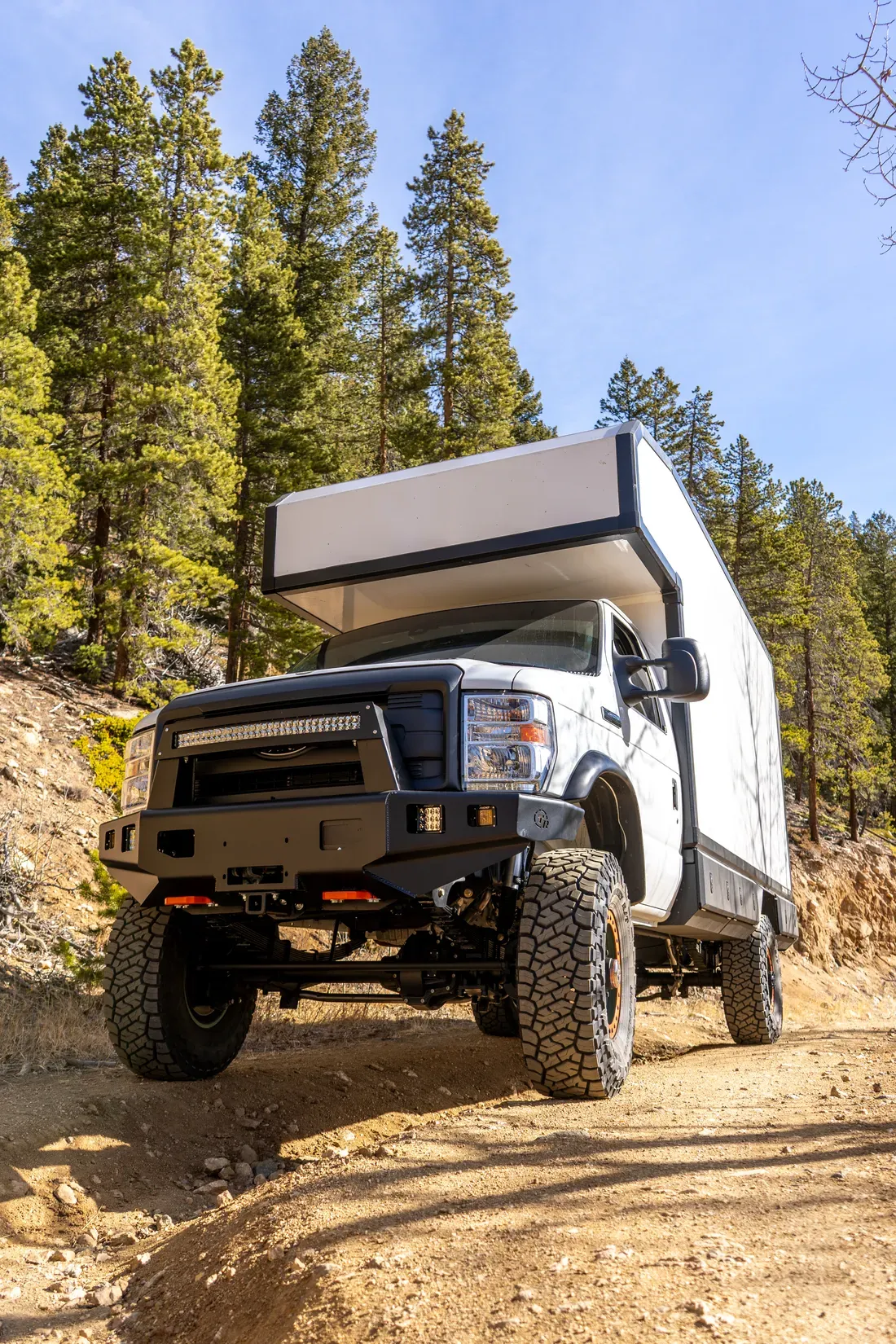 Ford E-Series 4x4 composite expedition camper climbing a rocky trail, front three-quarter view with the lifted suspension flexing over the terrain