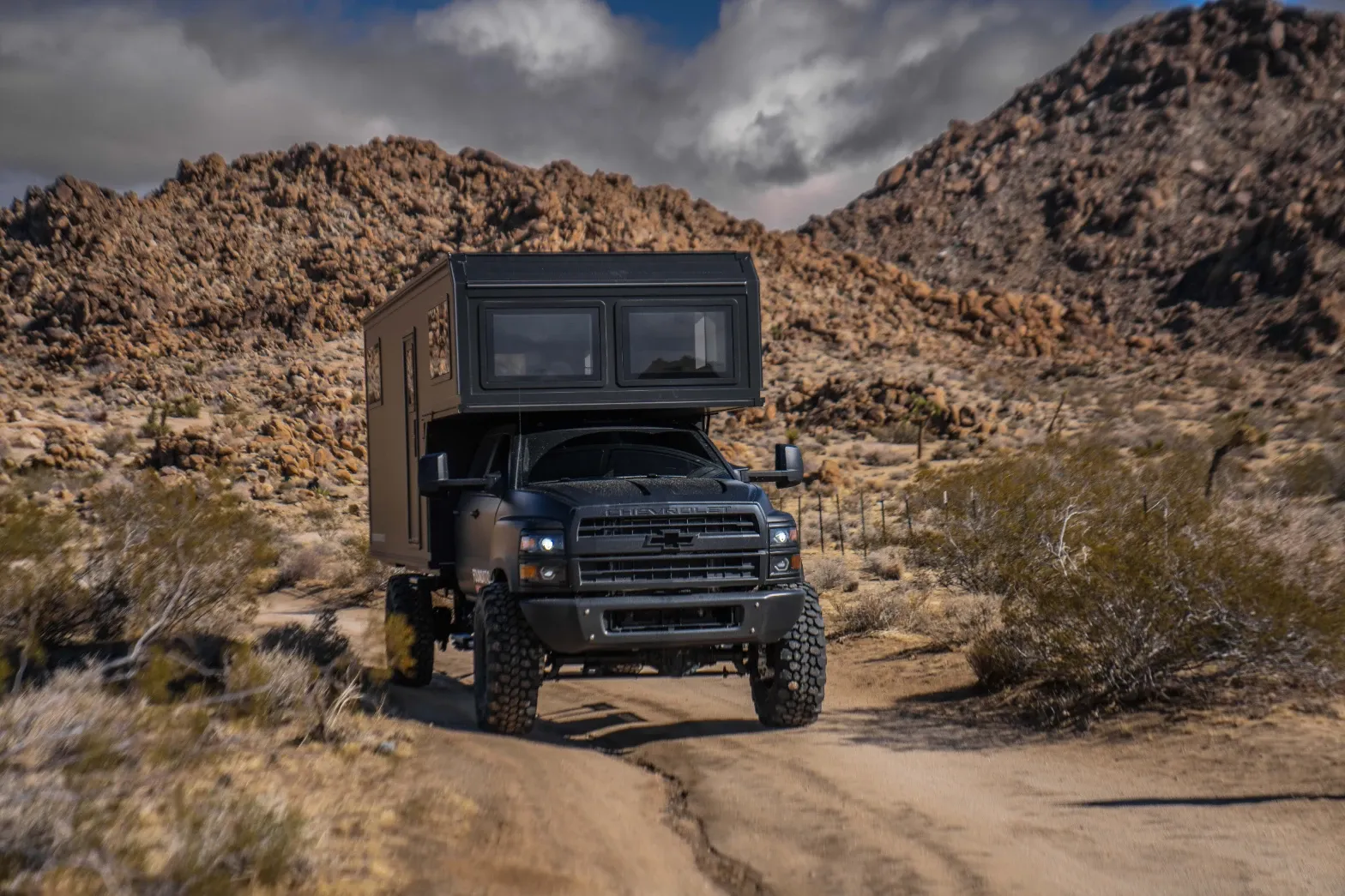 Blacked-out Chevrolet 6500 4x4 expedition camper on a desert trail below rocky hills