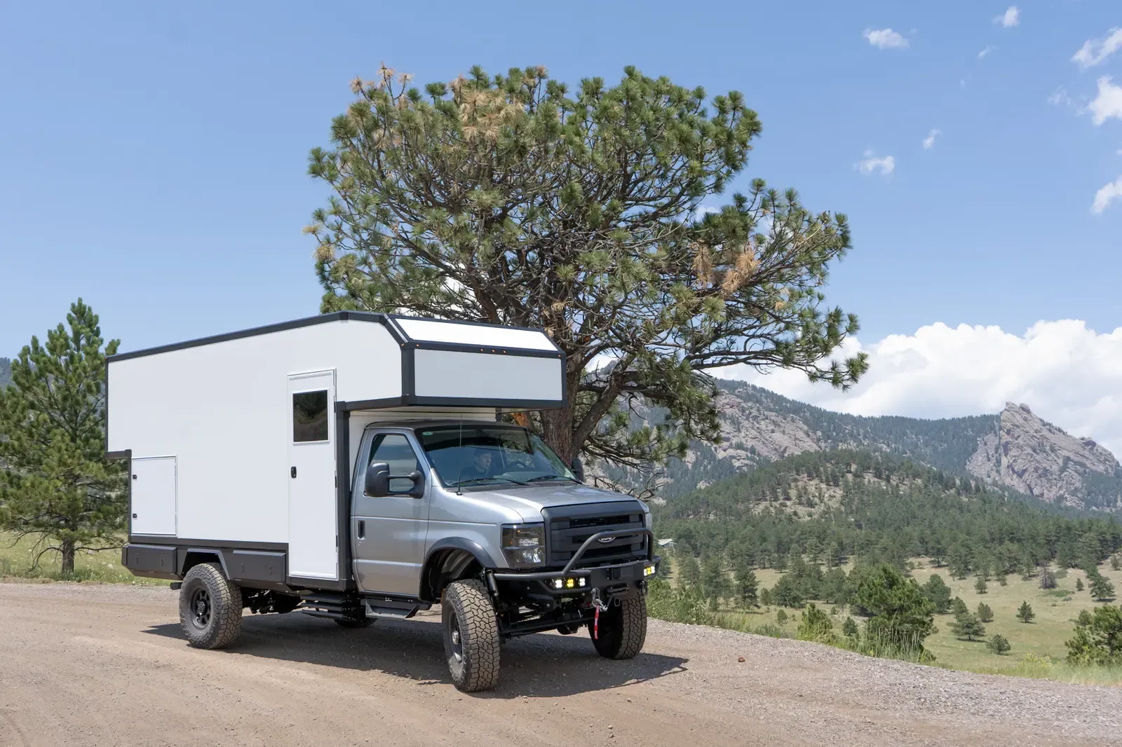 Silver-cab, white-cabover Ford E-450 expedition camper, side profile on a dirt road below the foothills