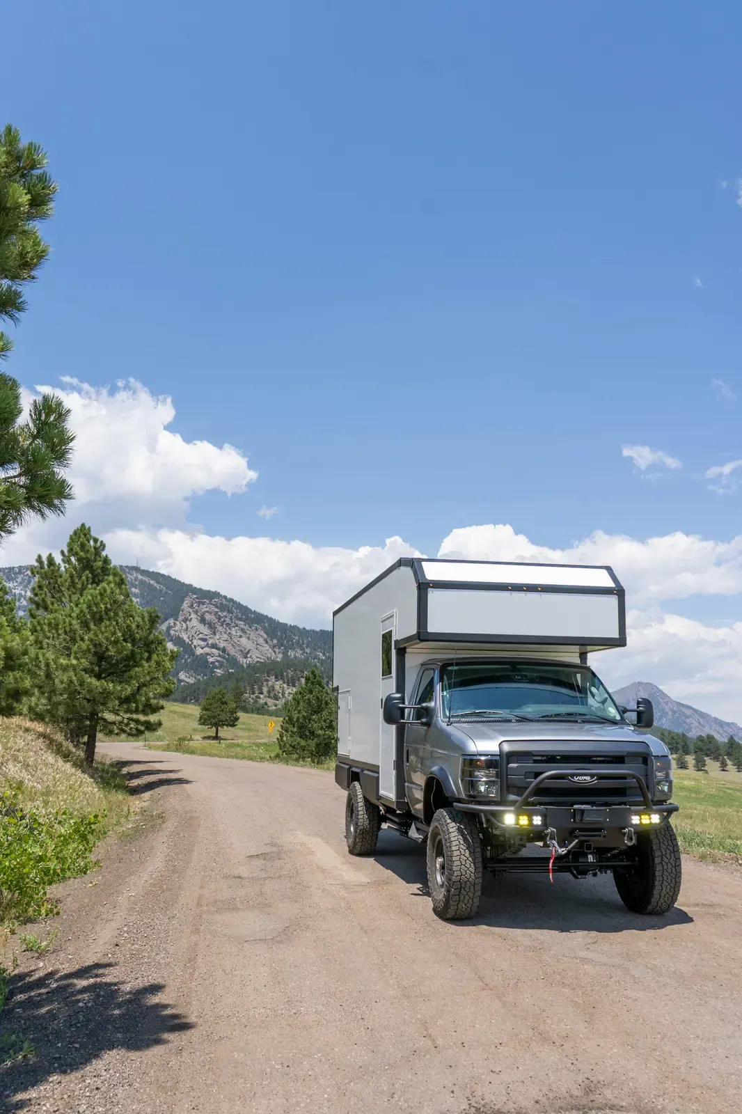 Silver-cab Ford E-450 expedition camper, front three-quarter view on a tree-lined dirt road