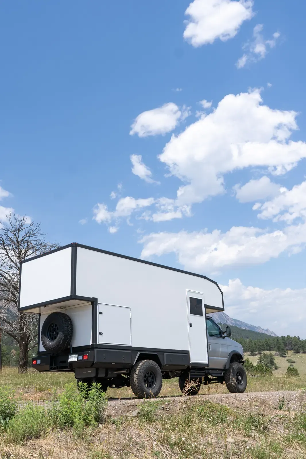 Silver-cab, white-cabover Ford E-450 expedition camper, rear three-quarter view with a spare tire on the back