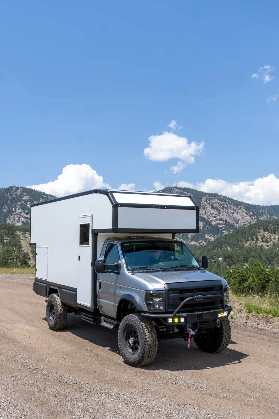 Silver-cab, white-cabover Ford E-450 expedition camper, front three-quarter view under a blue sky