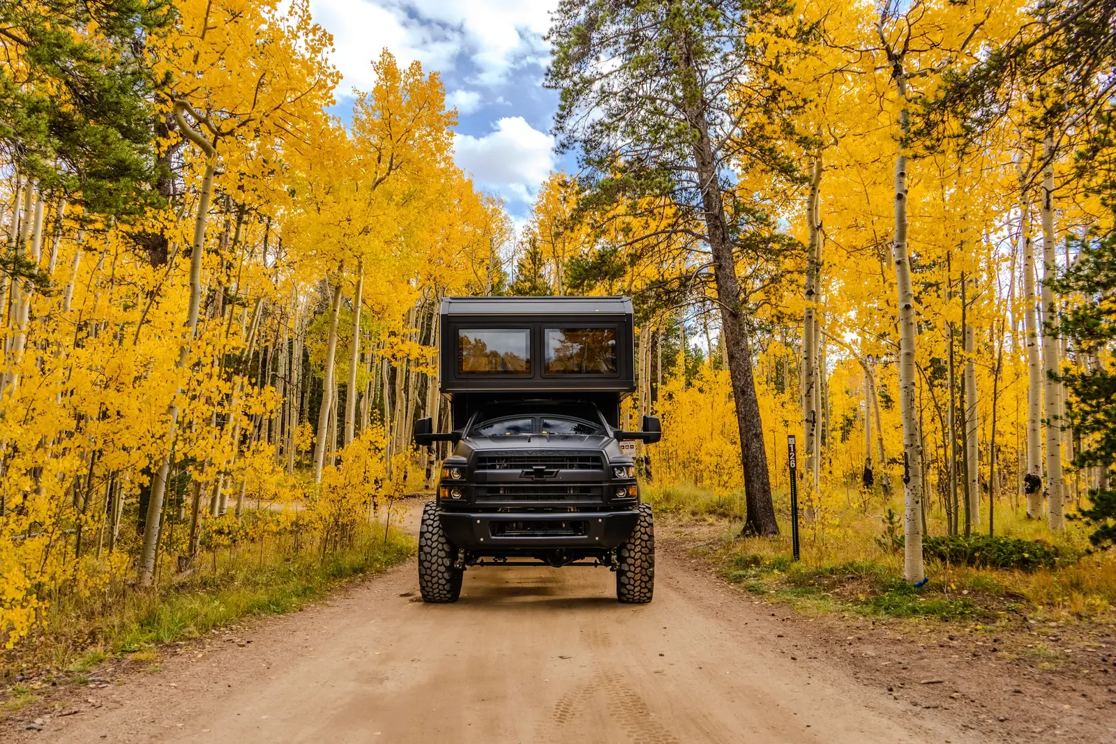 Blacked-out Chevrolet 6500 4x4 expedition camper, front three-quarter view on a dirt road framed by golden autumn aspens