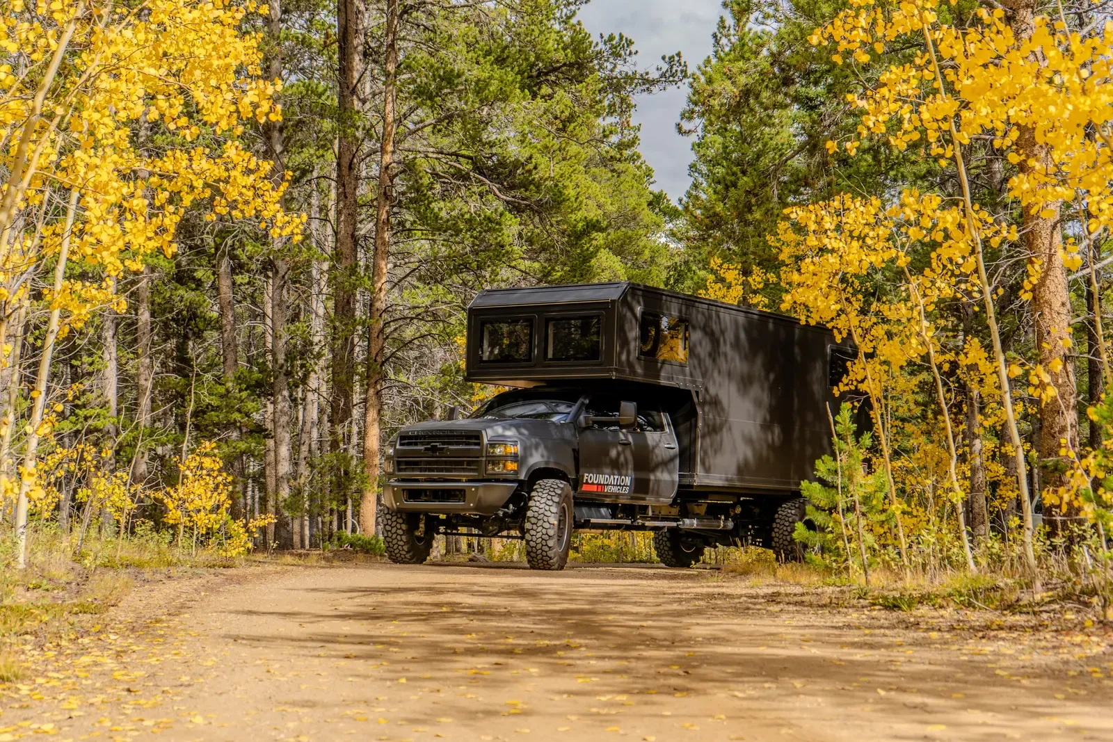 Blacked-out Chevrolet 6500 expedition camper, low front three-quarter view with sunlit aspen trunks behind