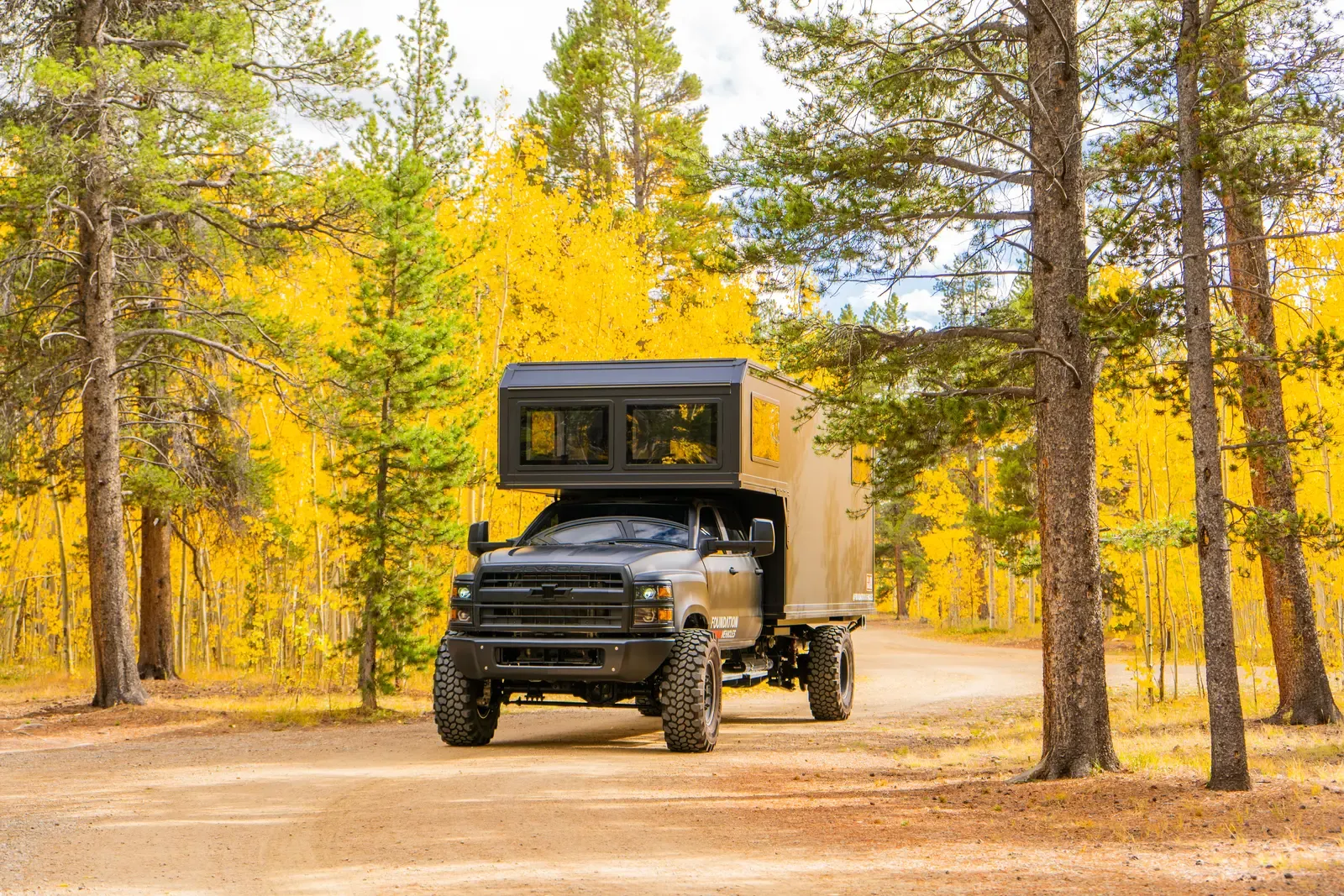 Blacked-out Chevrolet 6500 expedition camper on a leaf-strewn forest road beside golden aspens