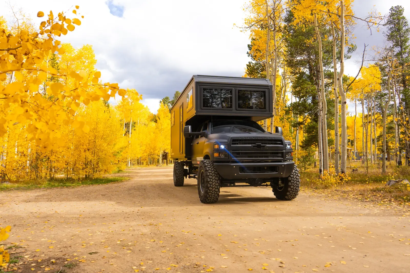 Blacked-out Chevrolet 6500 expedition camper, rear three-quarter view on a mountain road with golden foliage behind