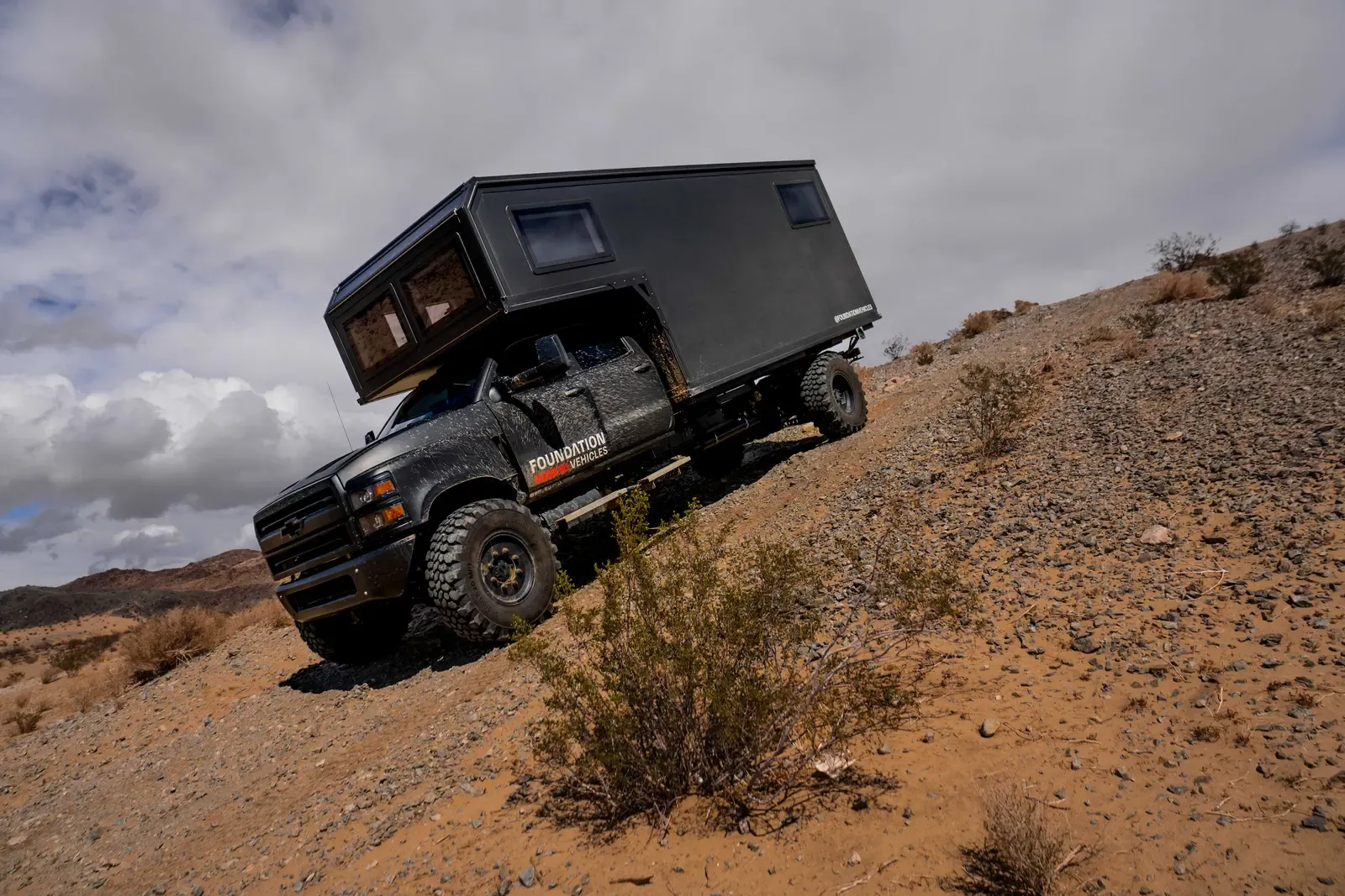 Blacked-out Chevrolet 6500 expedition camper climbing a steep desert embankment, side view