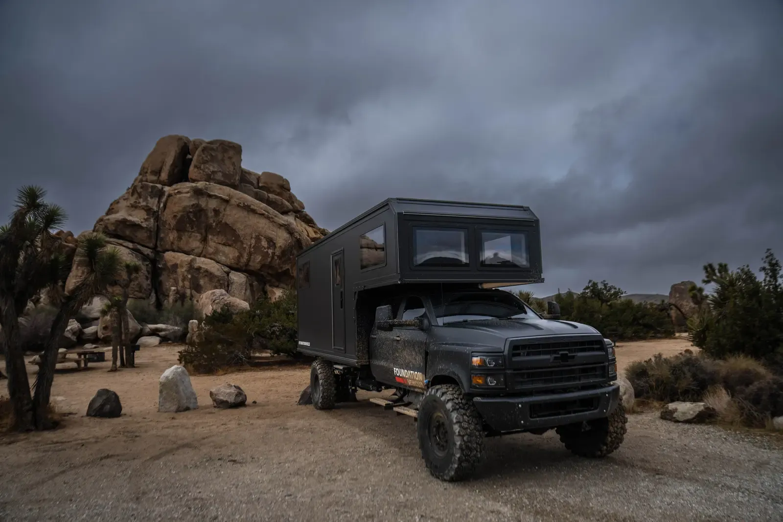 Blacked-out Chevrolet 6500 expedition camper among granite boulders, front three-quarter view under a moody sky