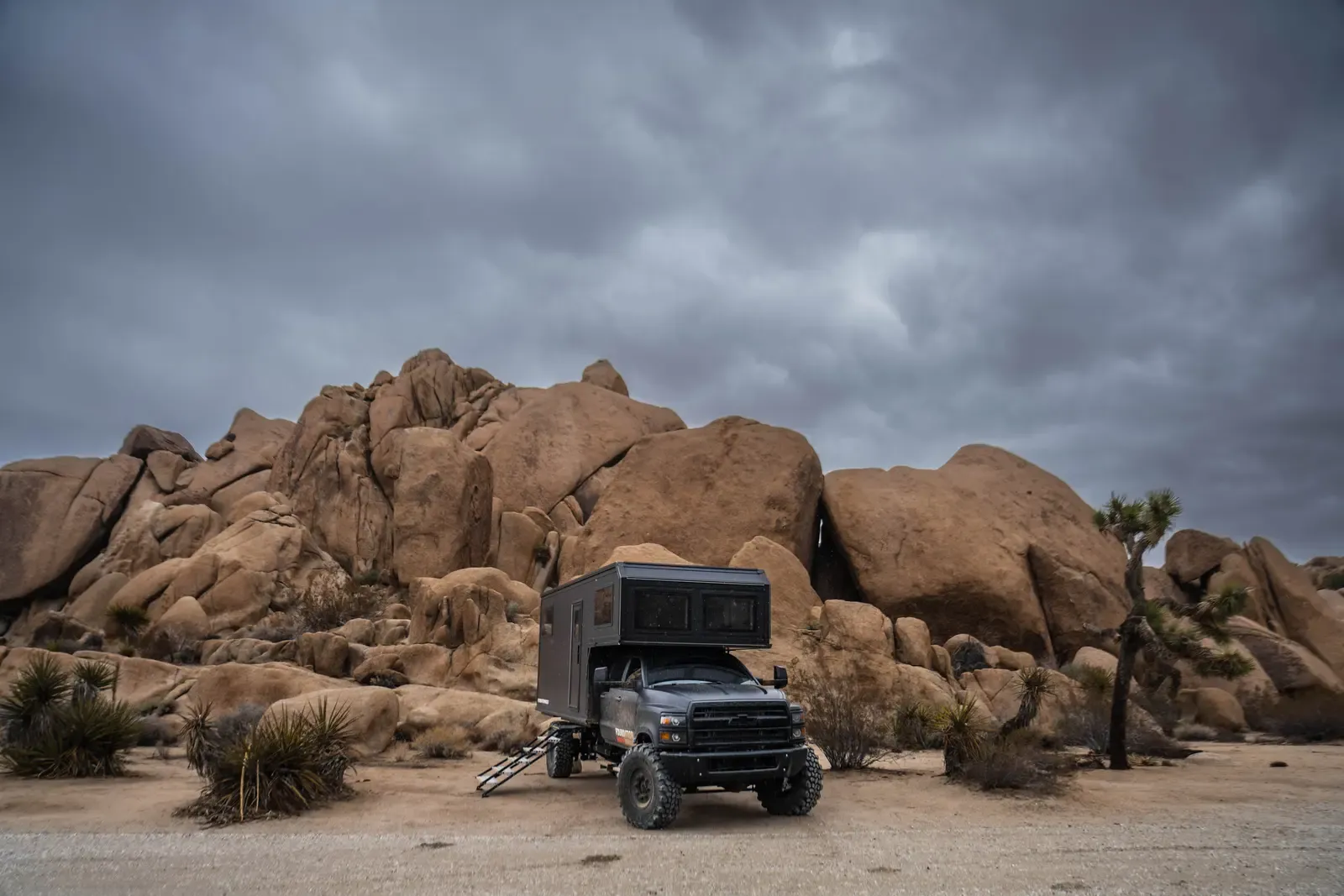 Blacked-out Chevrolet 6500 expedition camper among desert boulders, side view under a grey sky