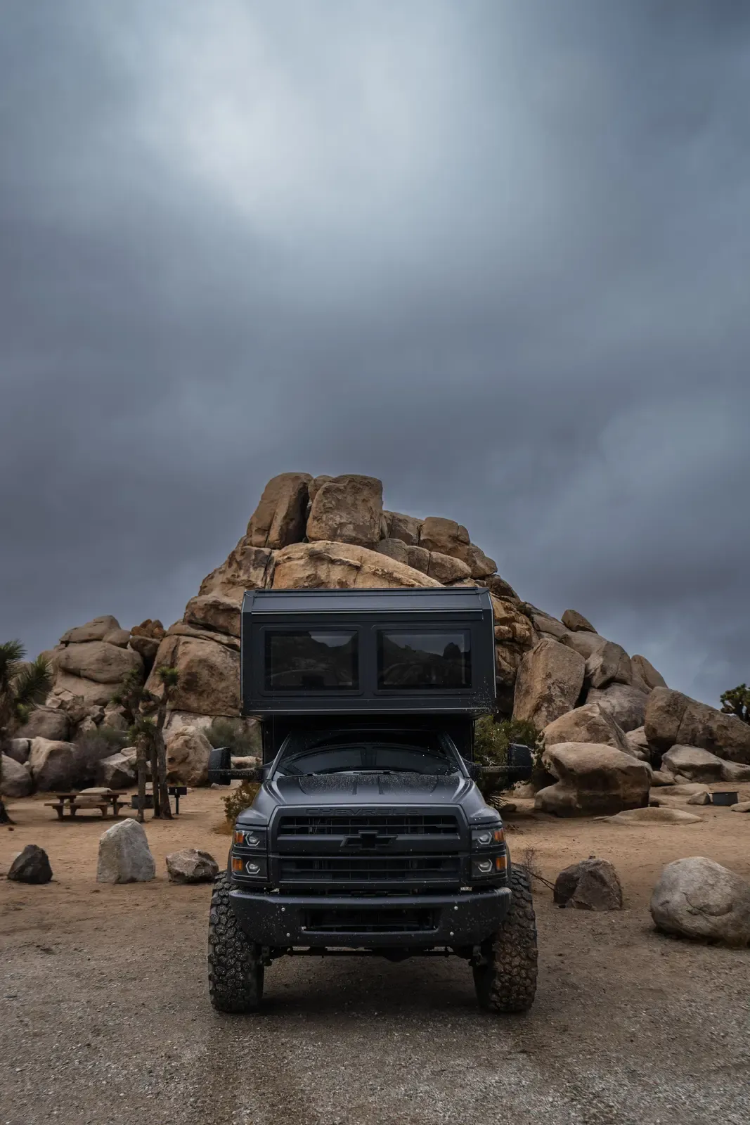 Blacked-out Chevrolet 6500 4x4 expedition camper on rocky desert ground, front three-quarter view
