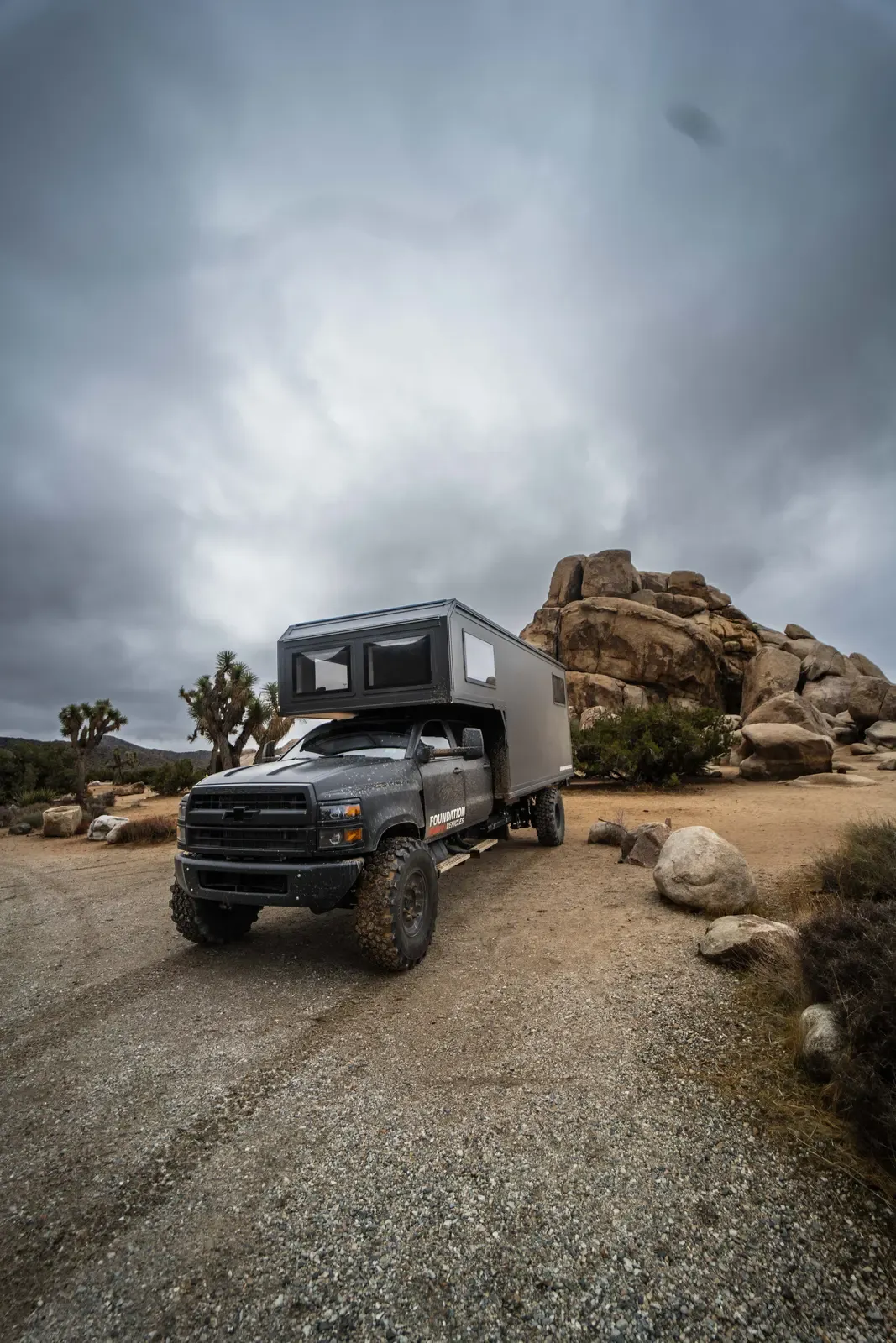 Blacked-out Chevrolet 6500 expedition camper on a desert gravel clearing under a heavy sky