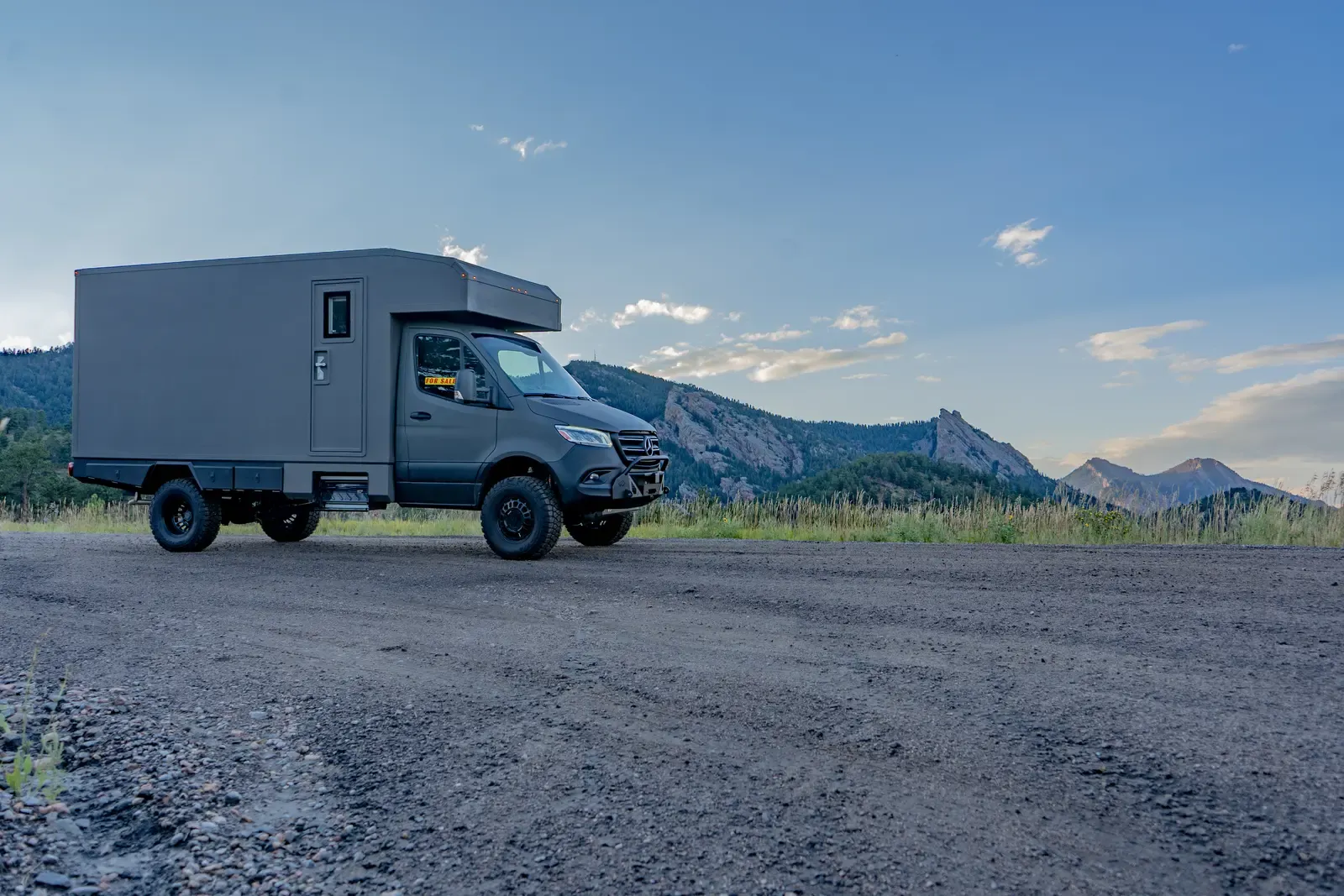 Matte-olive FV-170 demo composite camper on a Mercedes-Benz Sprinter 170, parked on an open gravel clearing below the mountains at dusk