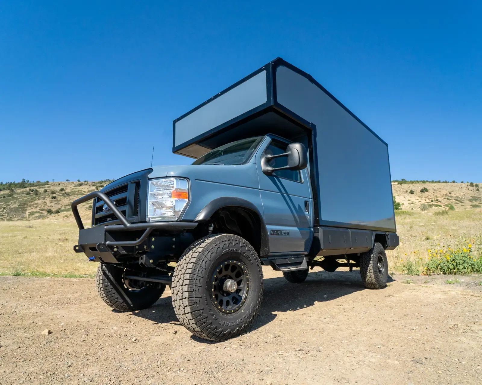 Cavalry Blue and white Ford E-Series E-158S 4x4 composite camper with black extrusions, front three-quarter view under a blue sky