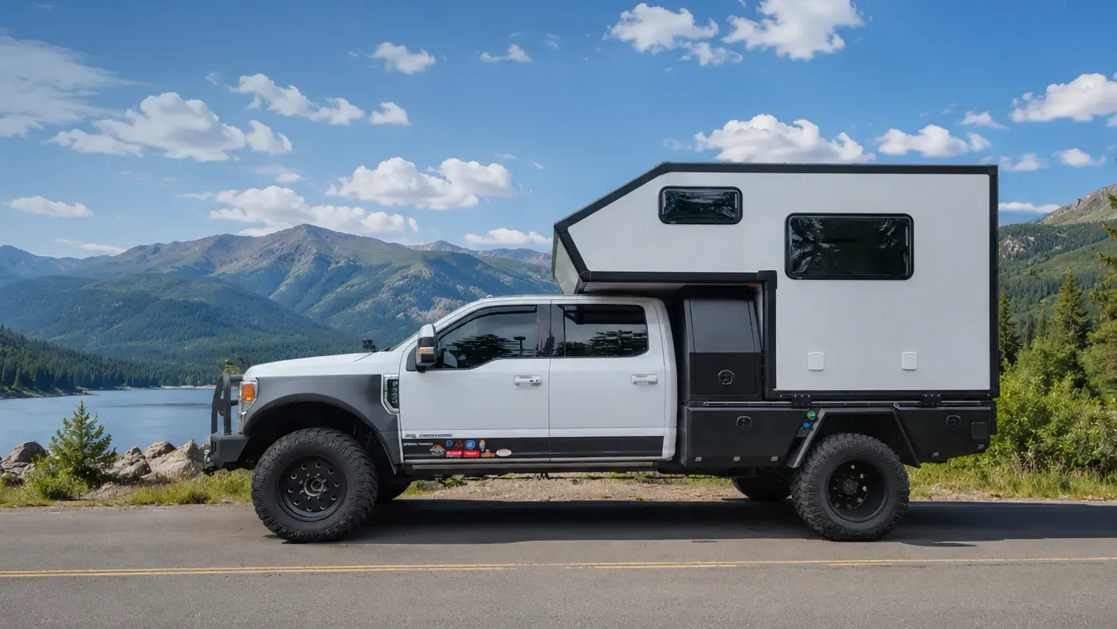 A composite flatbed camper on a Ford Super Duty flatbed truck, parked on a road beside a mountain lake