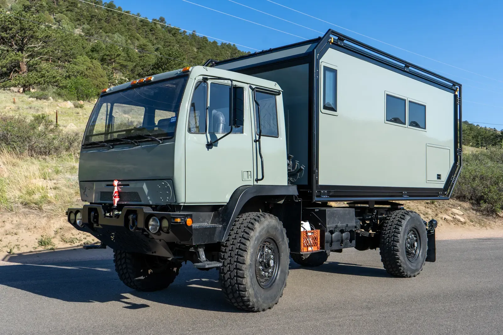 Sage-green LMTV expedition truck with a composite camper body, front three-quarter view with mountains behind