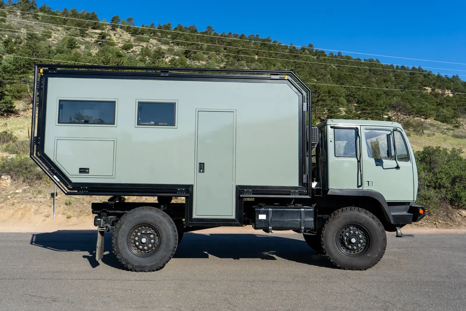 Sage-green LMTV expedition truck with a composite camper body, full-length side profile
