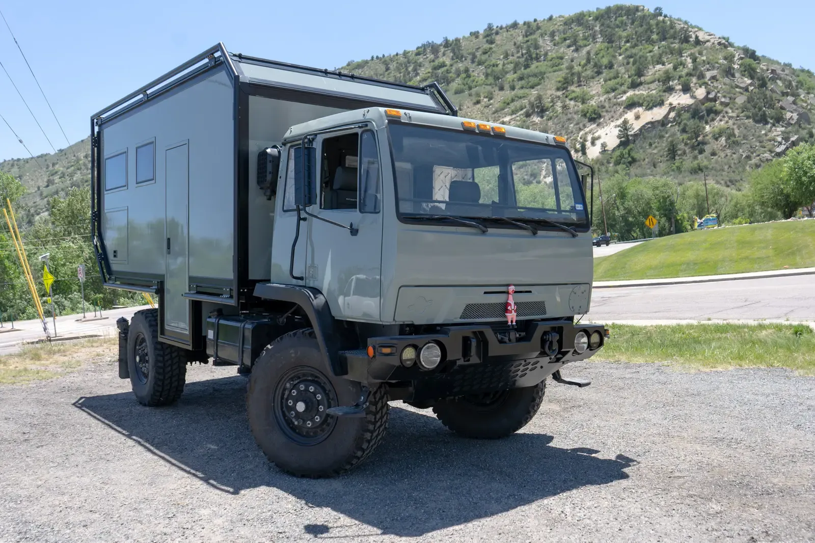 Sage-green LMTV expedition truck with a composite camper body, front three-quarter view below a wooded hill