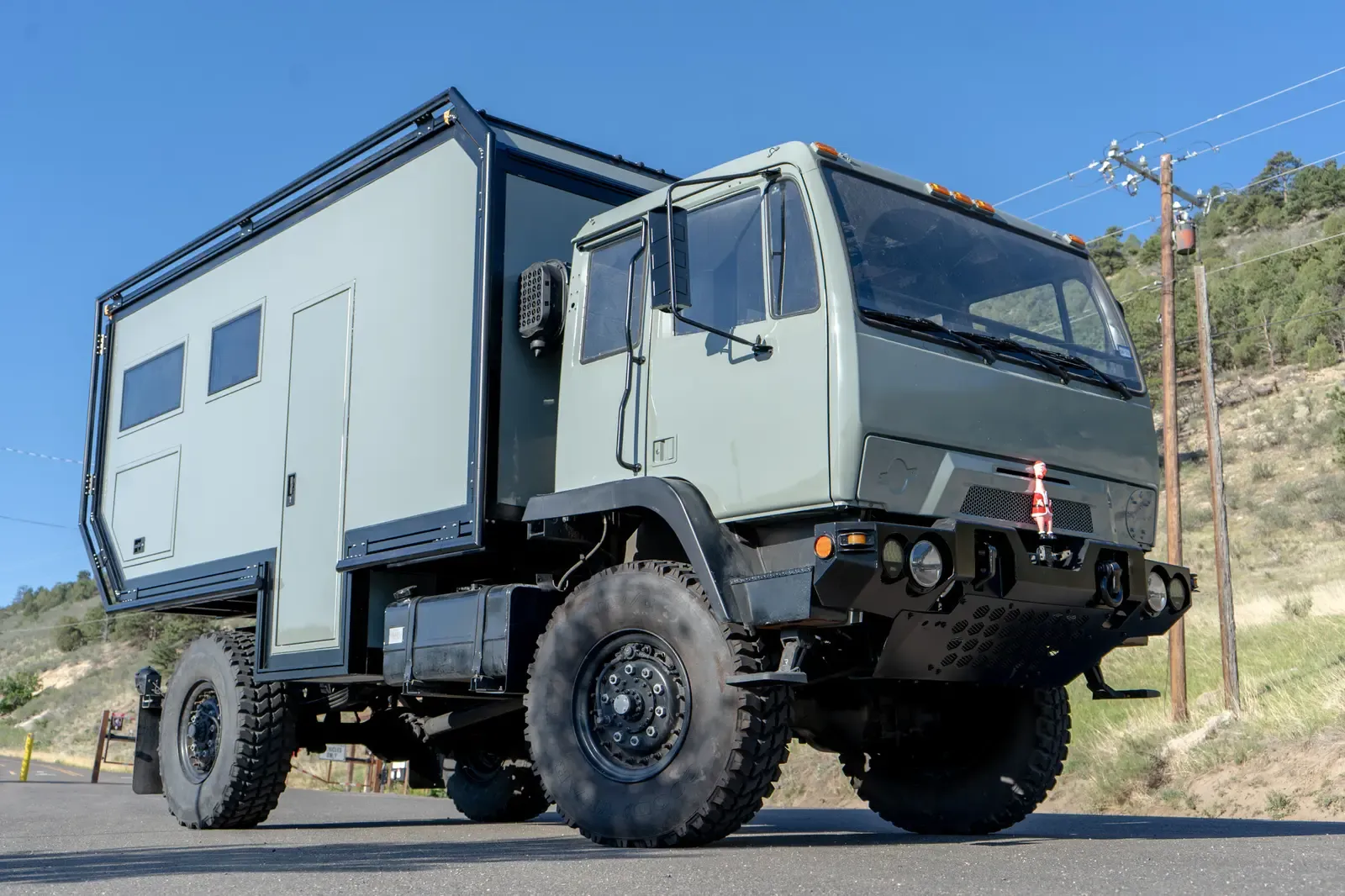 Sage-green LMTV expedition truck with a composite body, close front three-quarter view on its large off-road tires