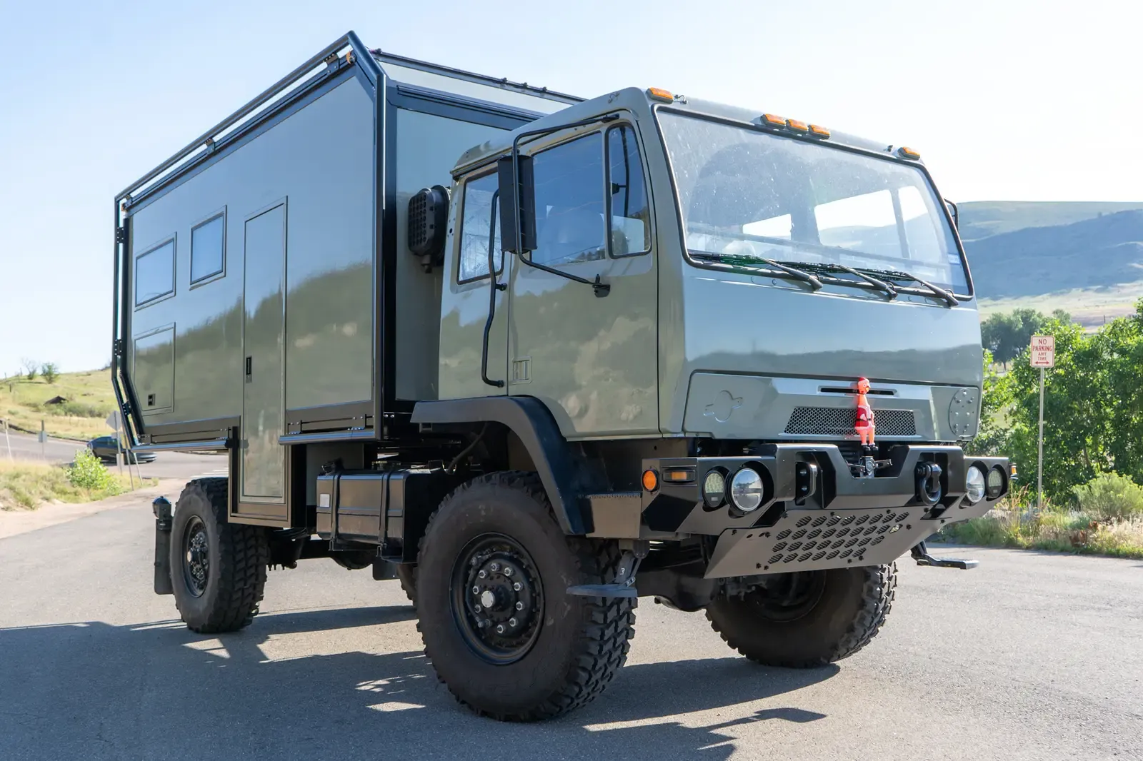 Sage-green LMTV expedition truck with a composite camper body, low front three-quarter view