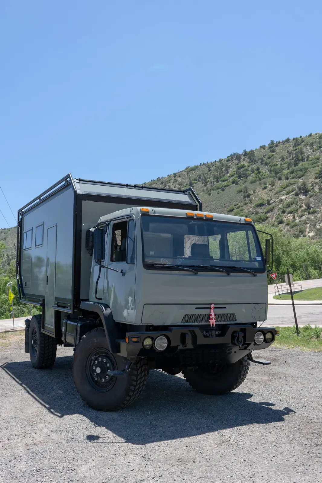 Sage-green LMTV expedition truck with a composite camper body at a distance on a paved road