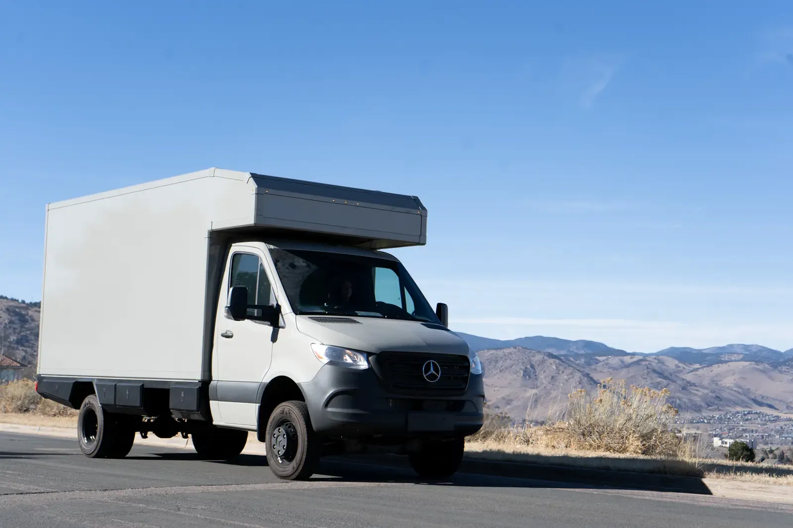 White commercial box body on a Mercedes-Benz Sprinter cab-chassis, front three-quarter view on a hilltop with mountains behind
