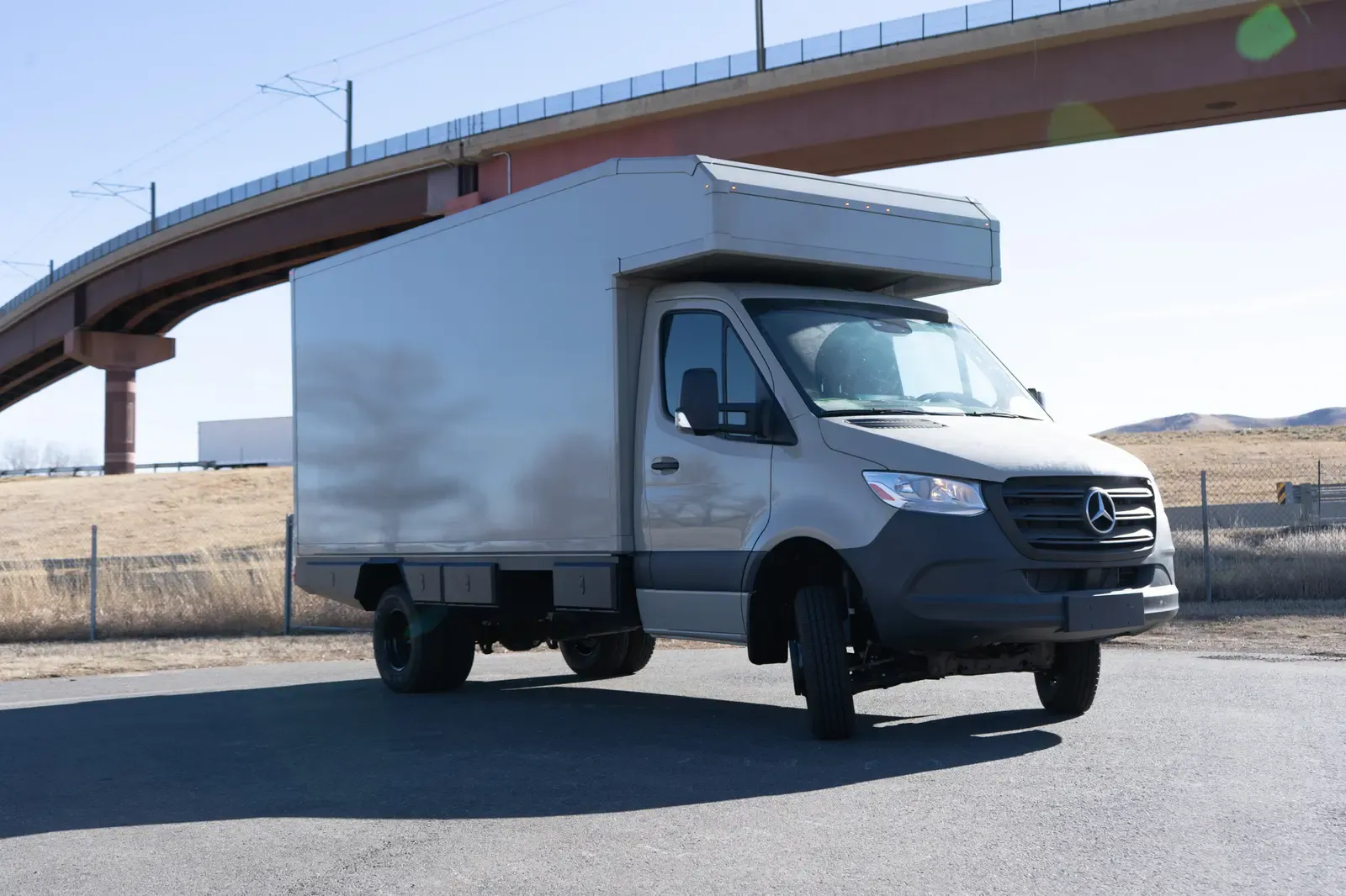 White commercial box body on a Mercedes-Benz Sprinter cab-chassis, front three-quarter view beneath a highway overpass