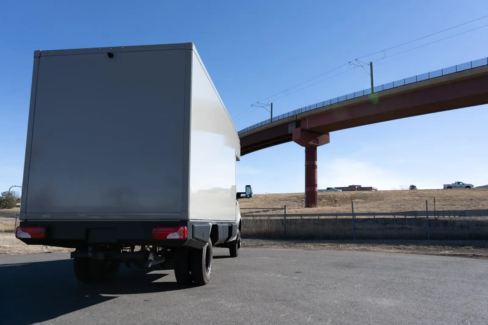 White commercial box body on a Mercedes-Benz Sprinter cab-chassis, rear three-quarter view beneath a highway overpass