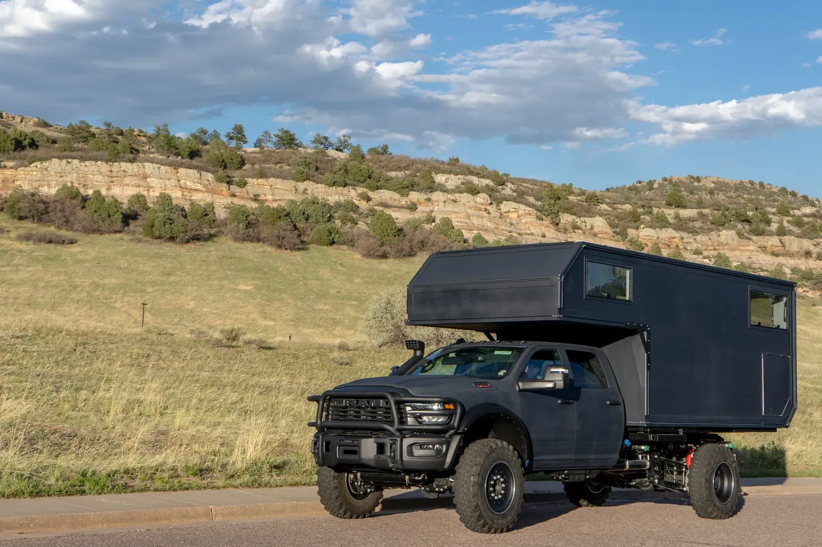 Black composite flatbed camper on a Ram 5500 cabover, front three-quarter view on a country road below grassy foothills