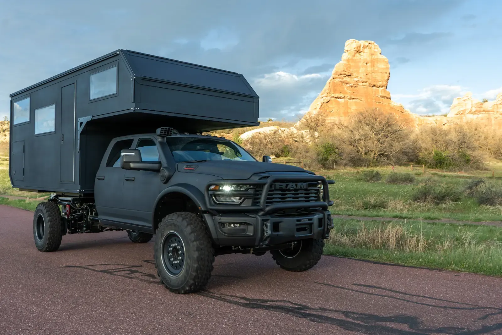 Black composite flatbed camper on a Ram 5500 cabover, front three-quarter view with a red sandstone formation behind