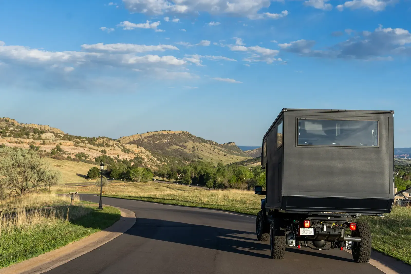 Black composite flatbed camper on a Ram 5500 cabover driving away down a country road, rear three-quarter view