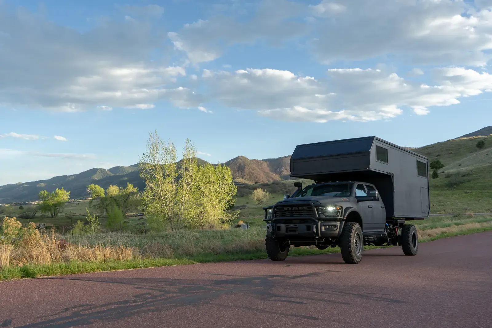Black composite flatbed camper on a Ram 5500 cabover, front three-quarter view on a mountain road in golden evening light
