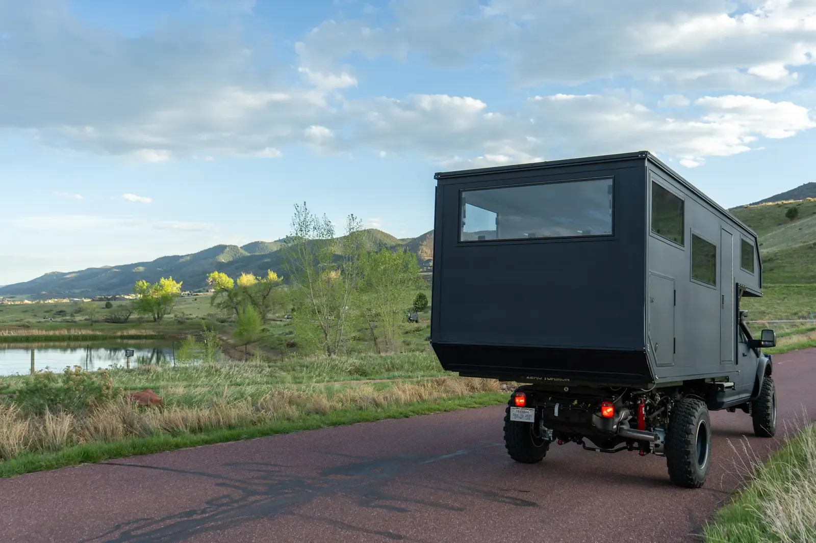 Black composite flatbed camper on a Ram 5500 cabover, rear three-quarter view on a road beside a lake