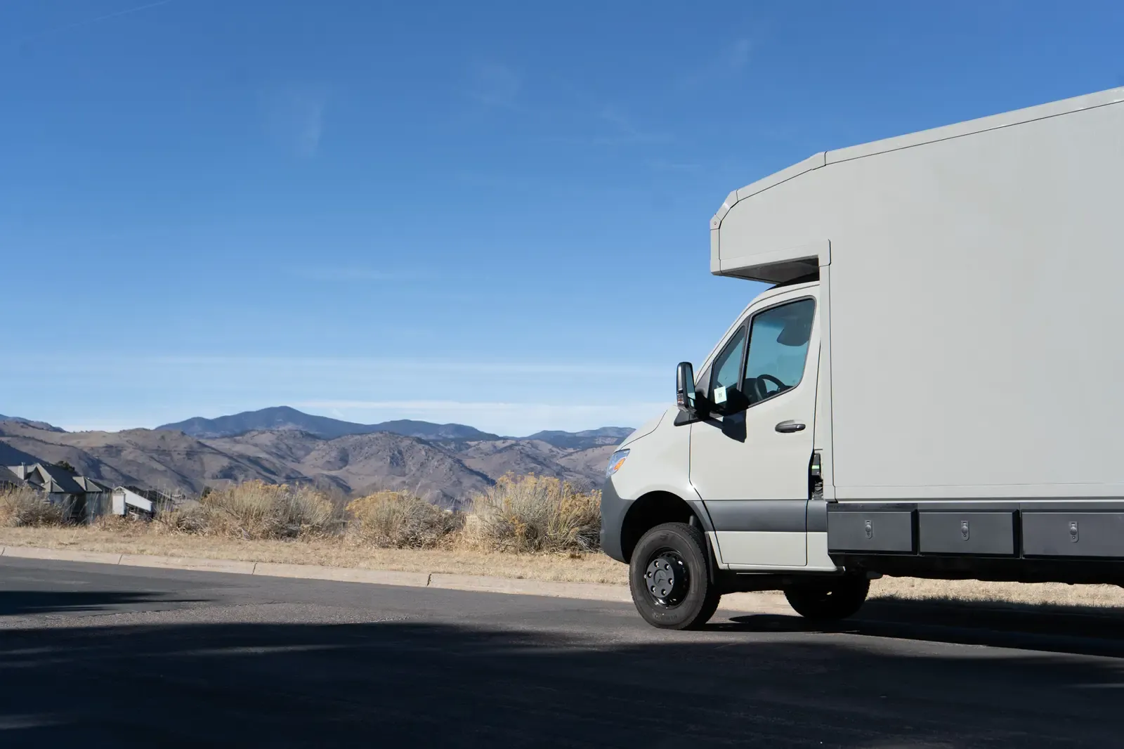 Stone grey commercial box body on a Mercedes-Benz Sprinter cab-chassis, front three-quarter view on a road with mountains