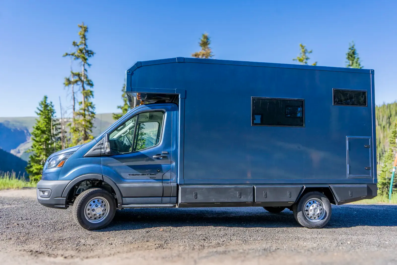 Blue composite box camper on a Ford Transit 156, front-side profile on a gravel pullout