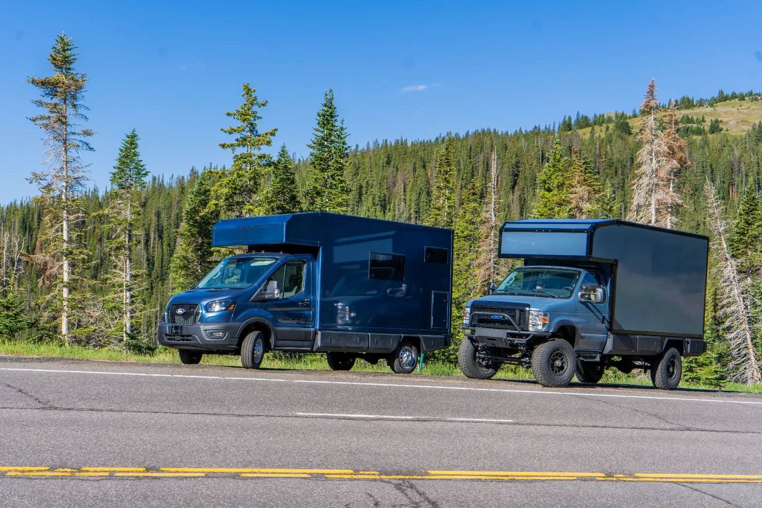 A Ford Transit 156 composite camper and a lifted Ford E-Series composite camper parked together on a forest highway