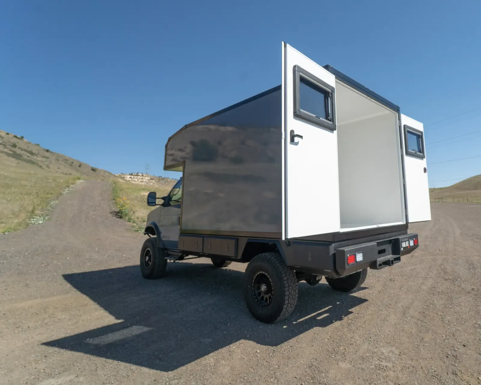 Two-tone composite box camper on a Ford E-Series 4x4, rear three-quarter view on a dirt road