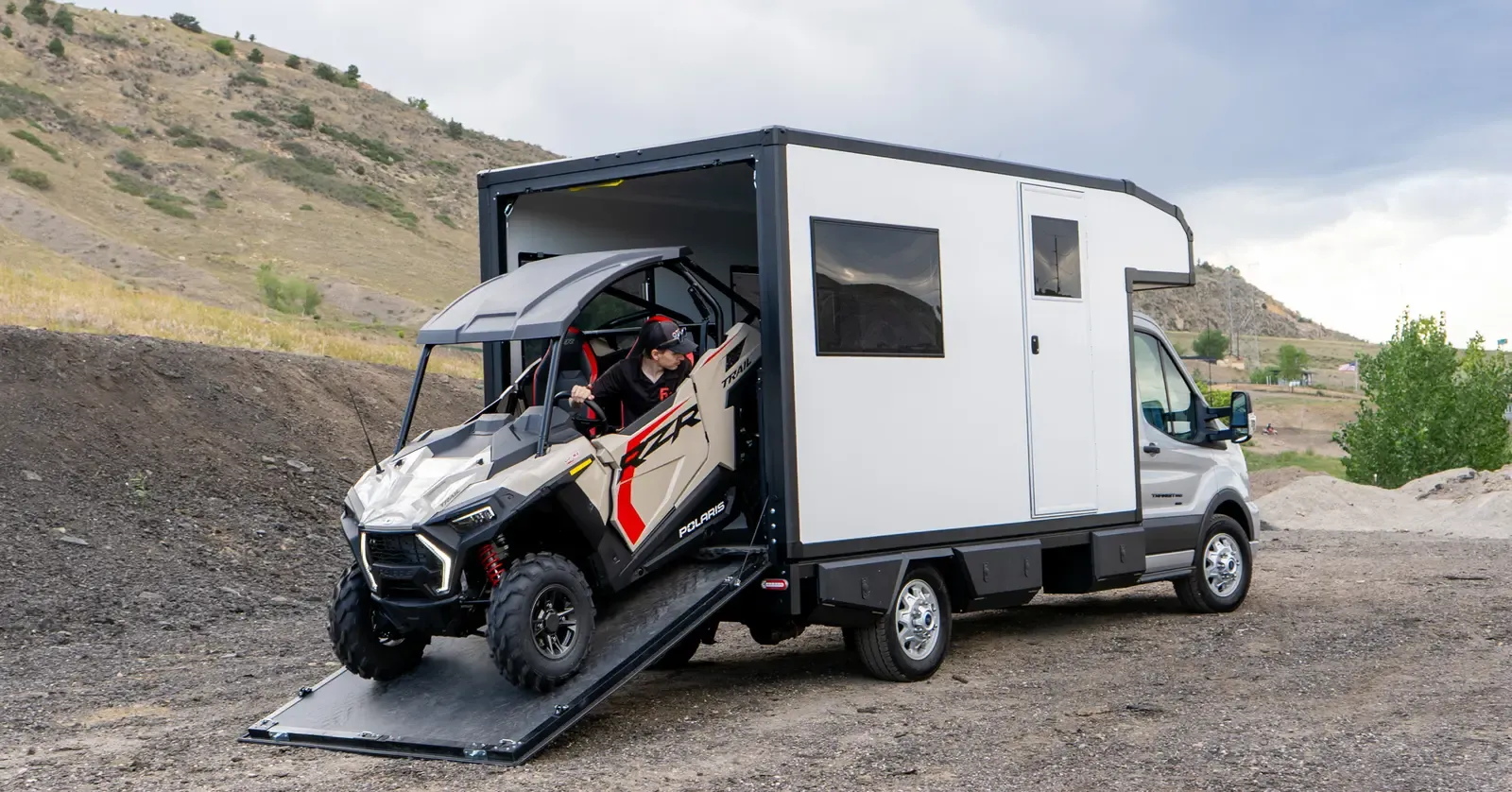 A side-by-side UTV parked on the ground beside a Foundation Vehicles RampDeck body on a Ford Transit 156, rear ramp down