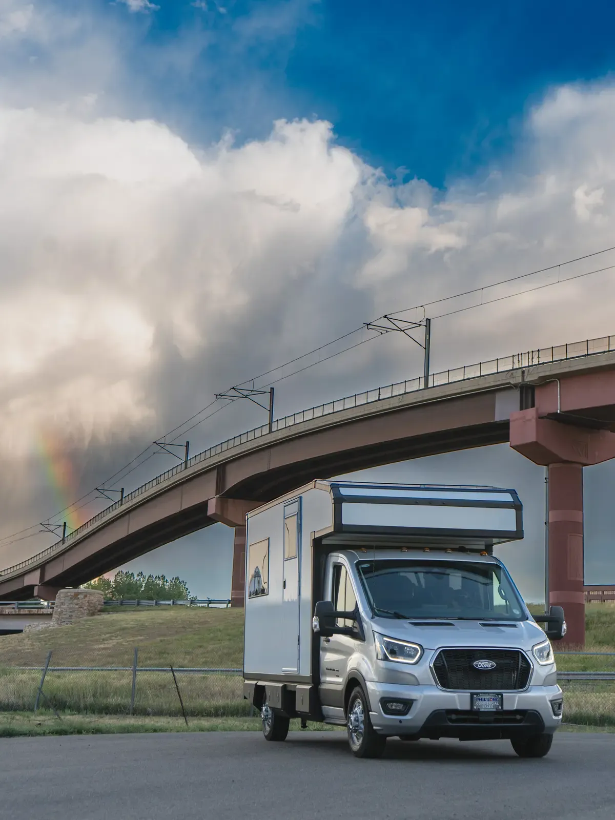 White Foundation Vehicles RampDeck body on a Ford Transit 156 driving on a highway beneath a dramatic storm sky with a rainbow