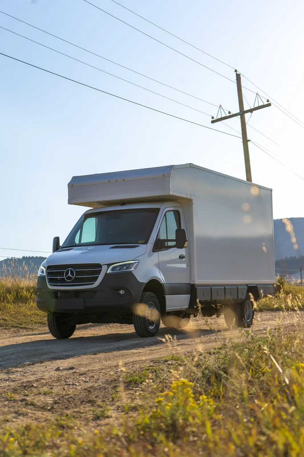 White composite box camper on a Mercedes-Benz Sprinter 170, front three-quarter view on a rural road below the hills