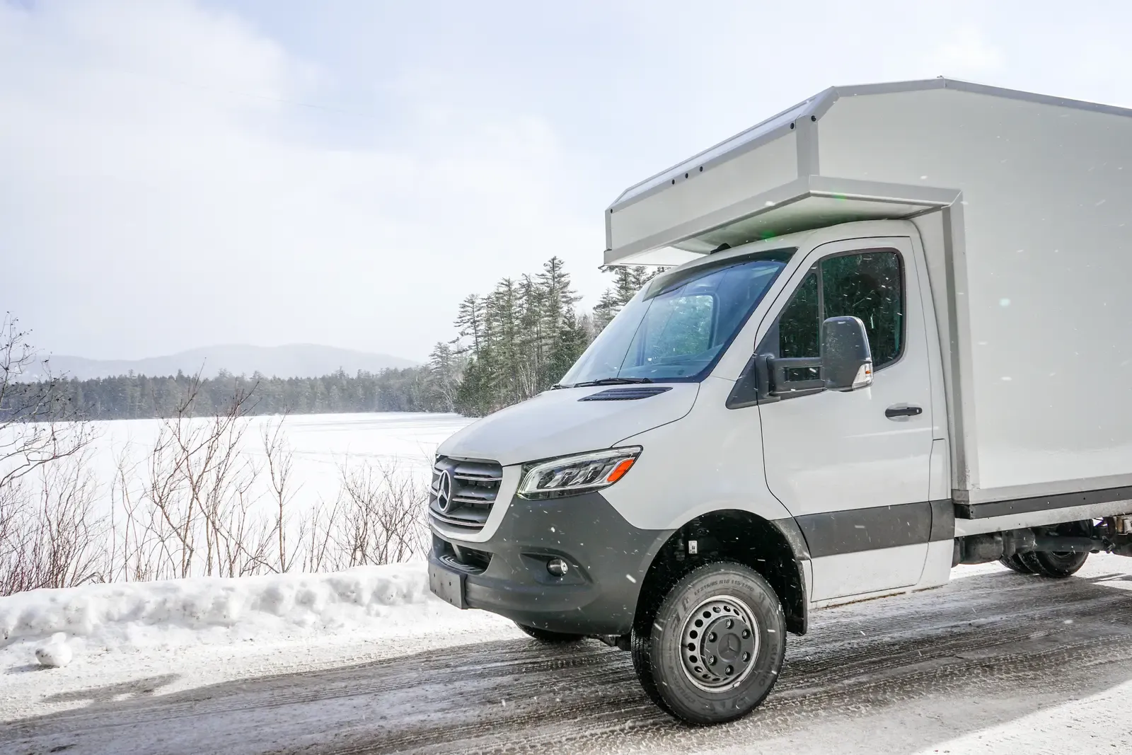 White composite box camper on an AWD Mercedes-Benz Sprinter 170, front three-quarter view beside a frozen lake in winter